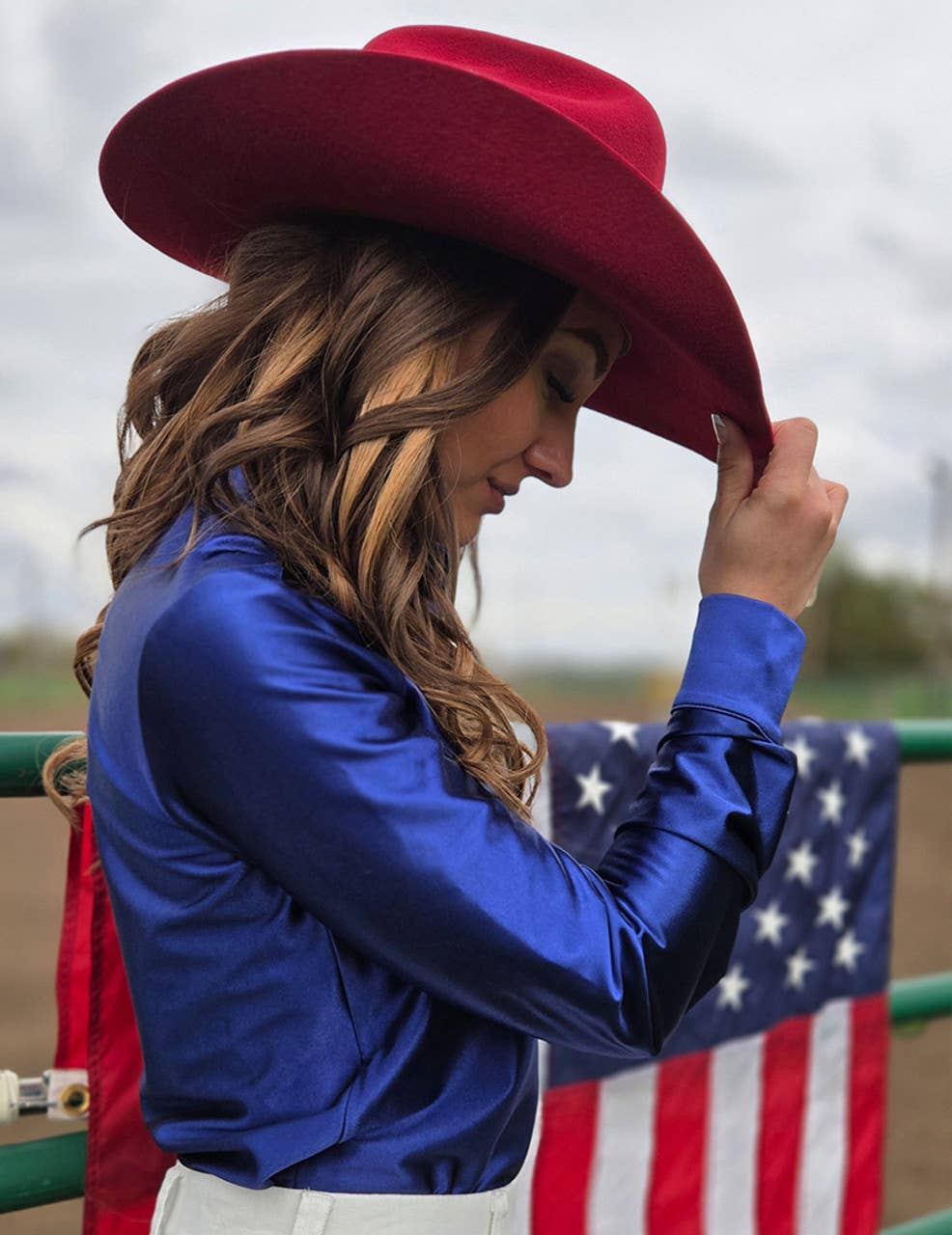 Cowgirl Tuff Rodeo Shirt indigo blue satin pullover styled with red hat and American flag background.