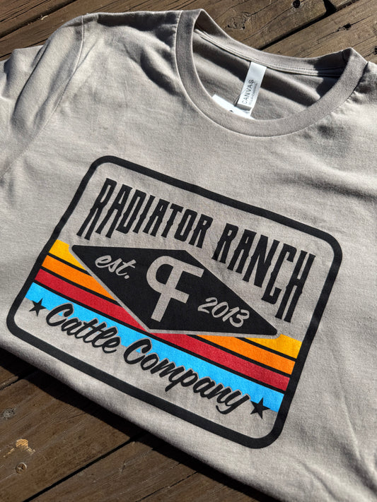A gray t-shirt with a colorful 'Radiator Ranch Cattle Company' serape-inspired print on the front, featuring text and geometric shapes.