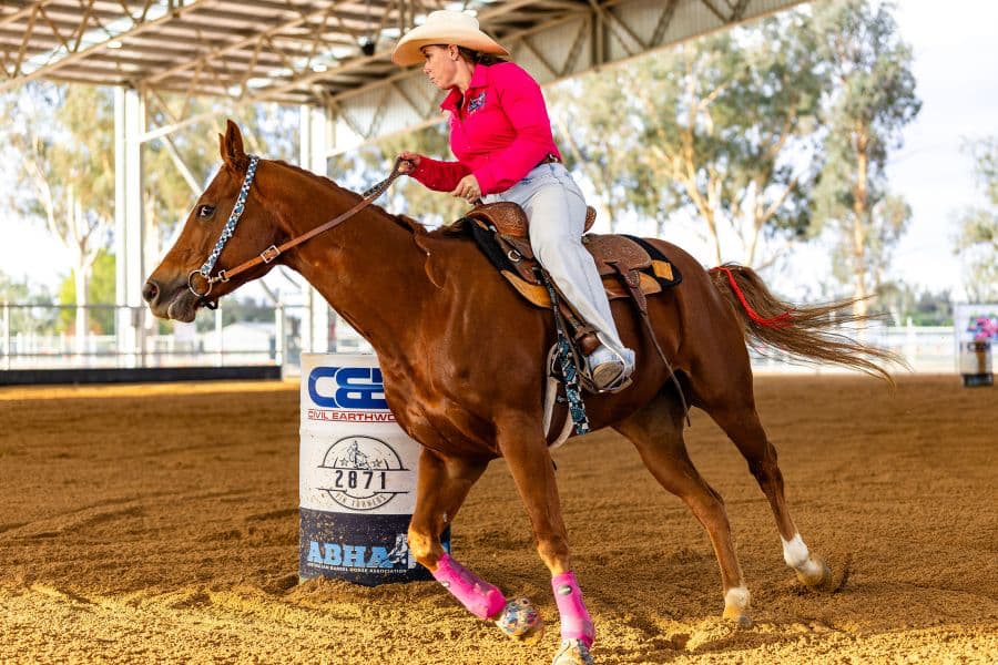 barrel racer in a pink shirt riding a brown horse around a barrel at an indoor arena, wearing custom western tack from bar3H Equine.