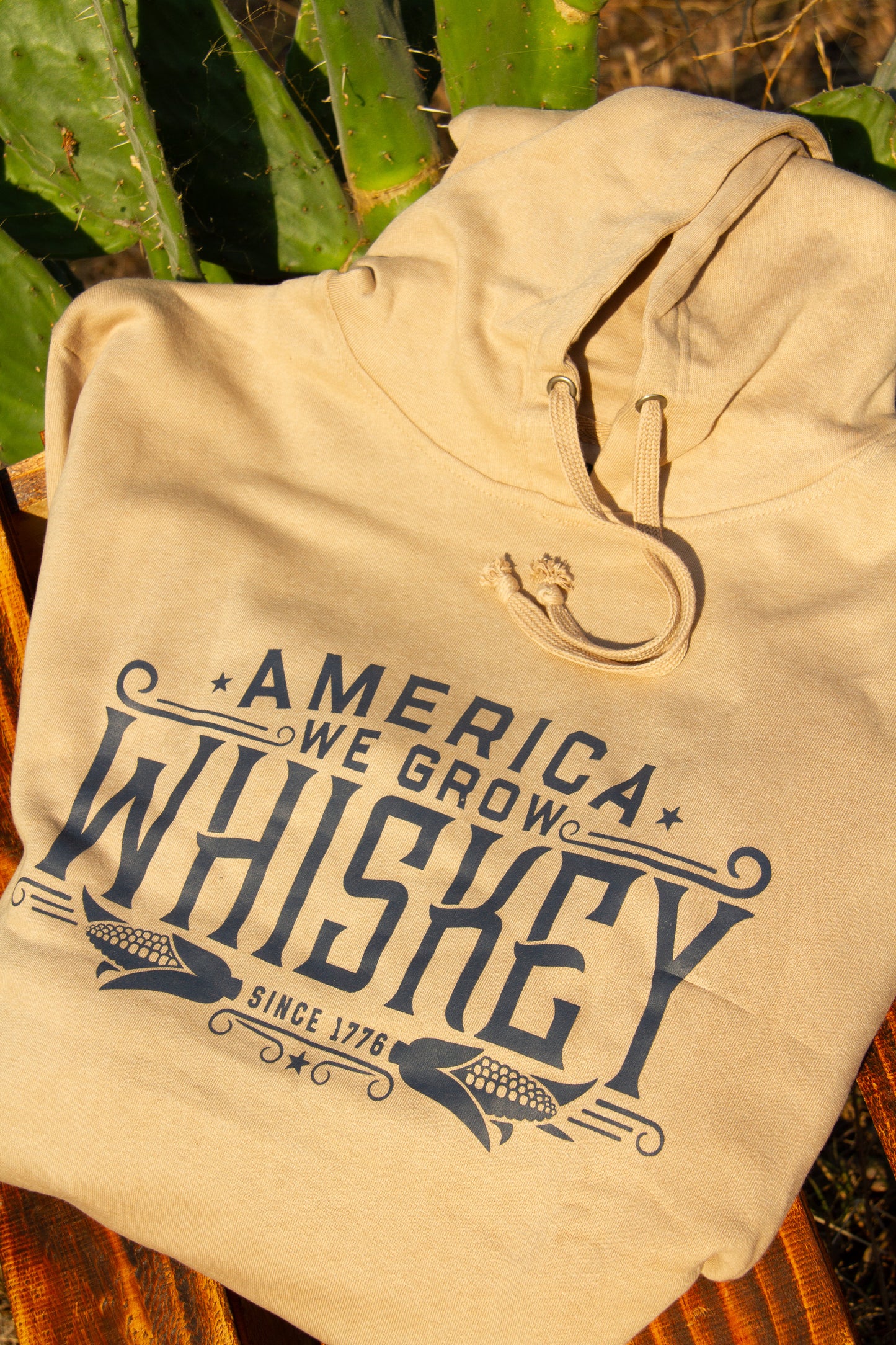 Beige hoodie with 'America We Grow Whiskey' text on a wooden surface with greenery in the background