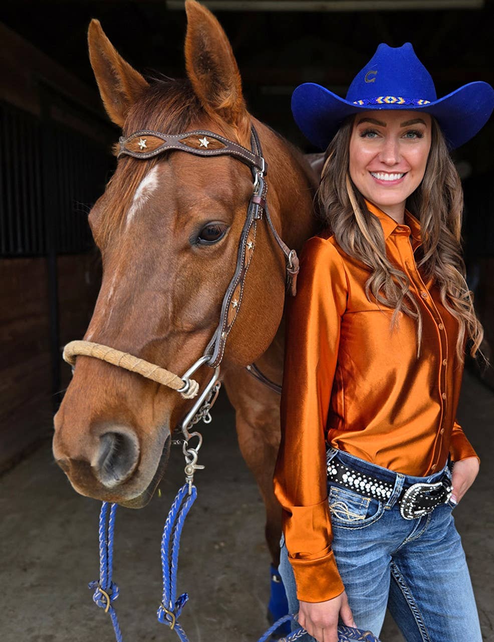 Cowgirl Tuff Rodeo Shirt Copper Satin Pullover worn by a smiling woman beside a brown horse in a barn.