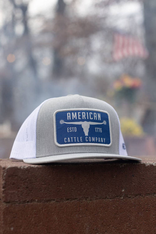 Gray and white cap with 'American Cattle Company' logo on a brick wall.