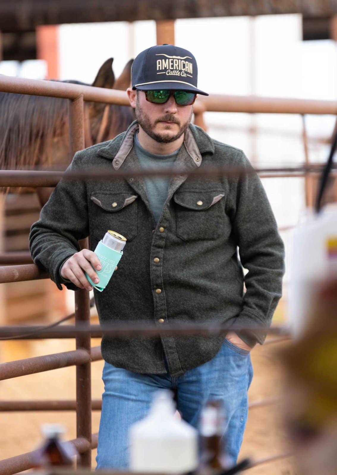 Man wearing a cap and sunglasses, holding a can, standing next to a horse in a barn.