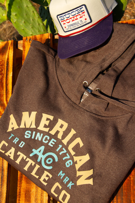 Grey hoodie with 'American Cattle Co' text and a cap on a wooden surface