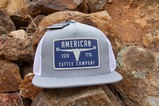 Gray and white cap with 'American Cattle Company' logo on a rocky surface