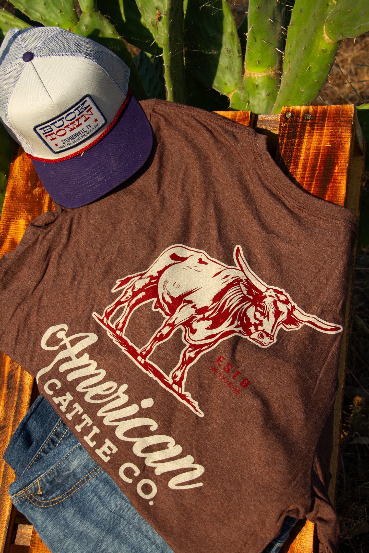 Brown t-shirt with red bull graphic and 'American Cattle Co.' text, paired with a cap, against a natural background.
