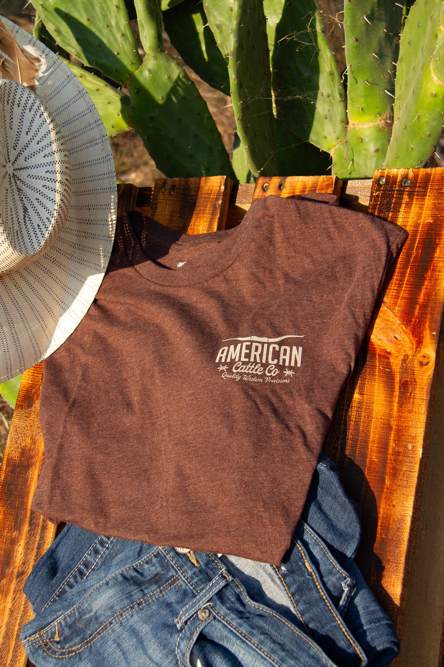 Brown t-shirt with 'American Cattle Co' logo on a wooden surface with cacti in the background