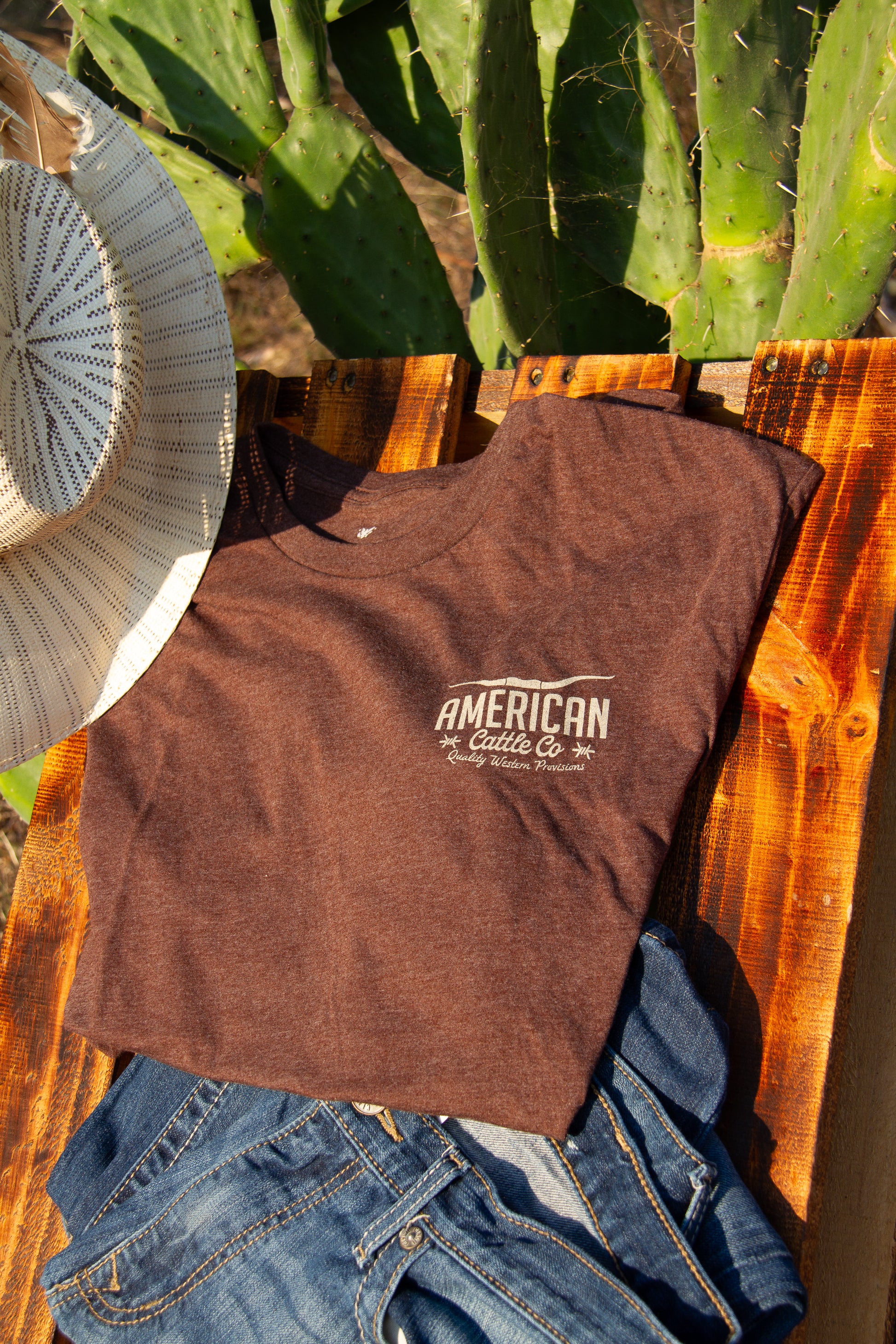Brown t-shirt with 'American Cattle Co' logo on a wooden surface with cacti in the background