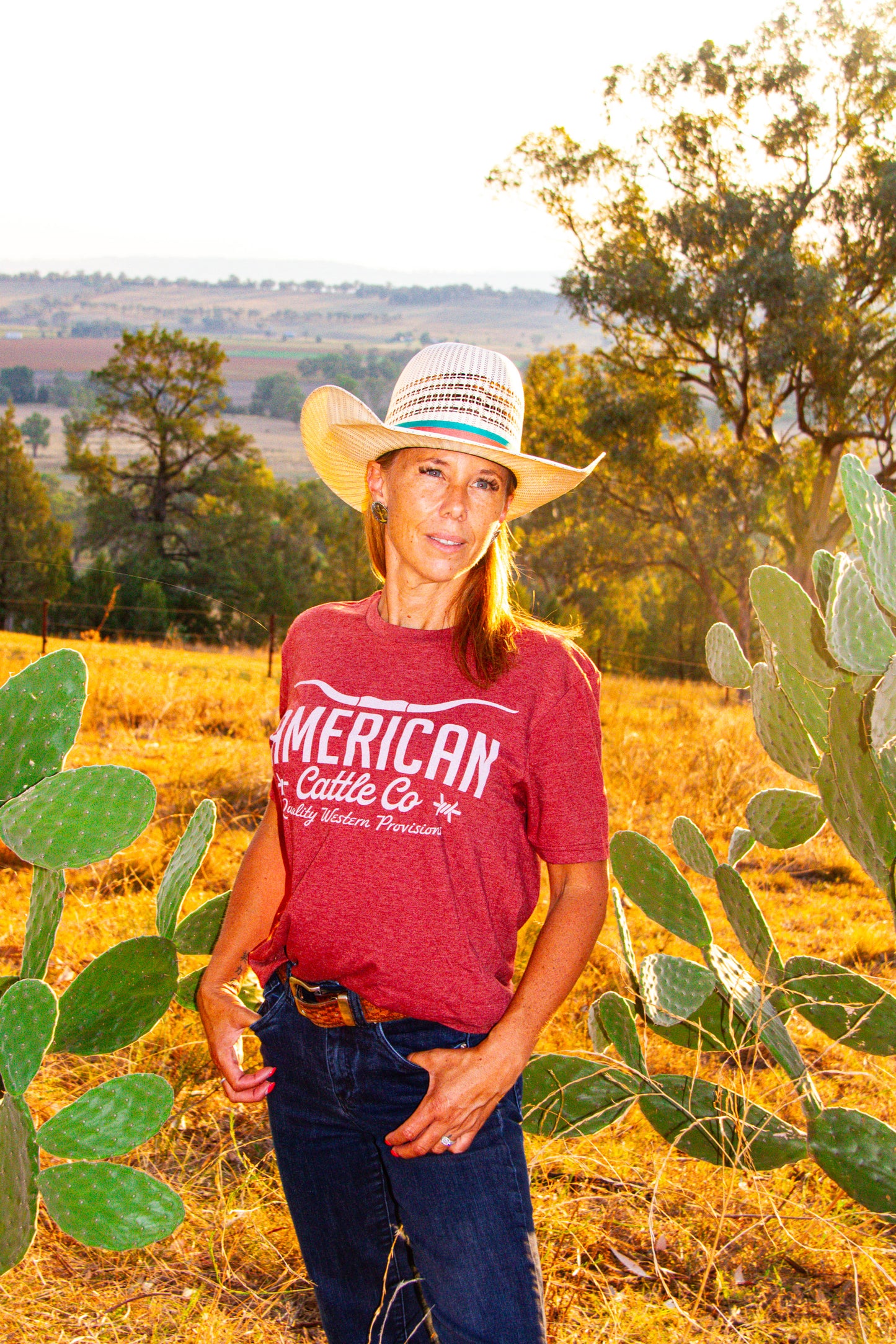Person wearing a red 'American Cattle Co' shirt standing in a field with cacti and trees.