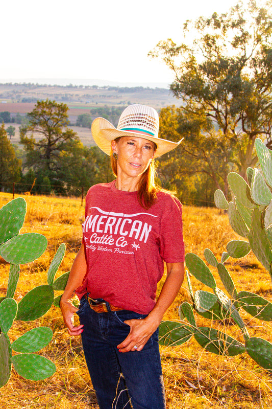 Person wearing a red 'American Cattle Co' shirt standing in a field with cacti and trees.