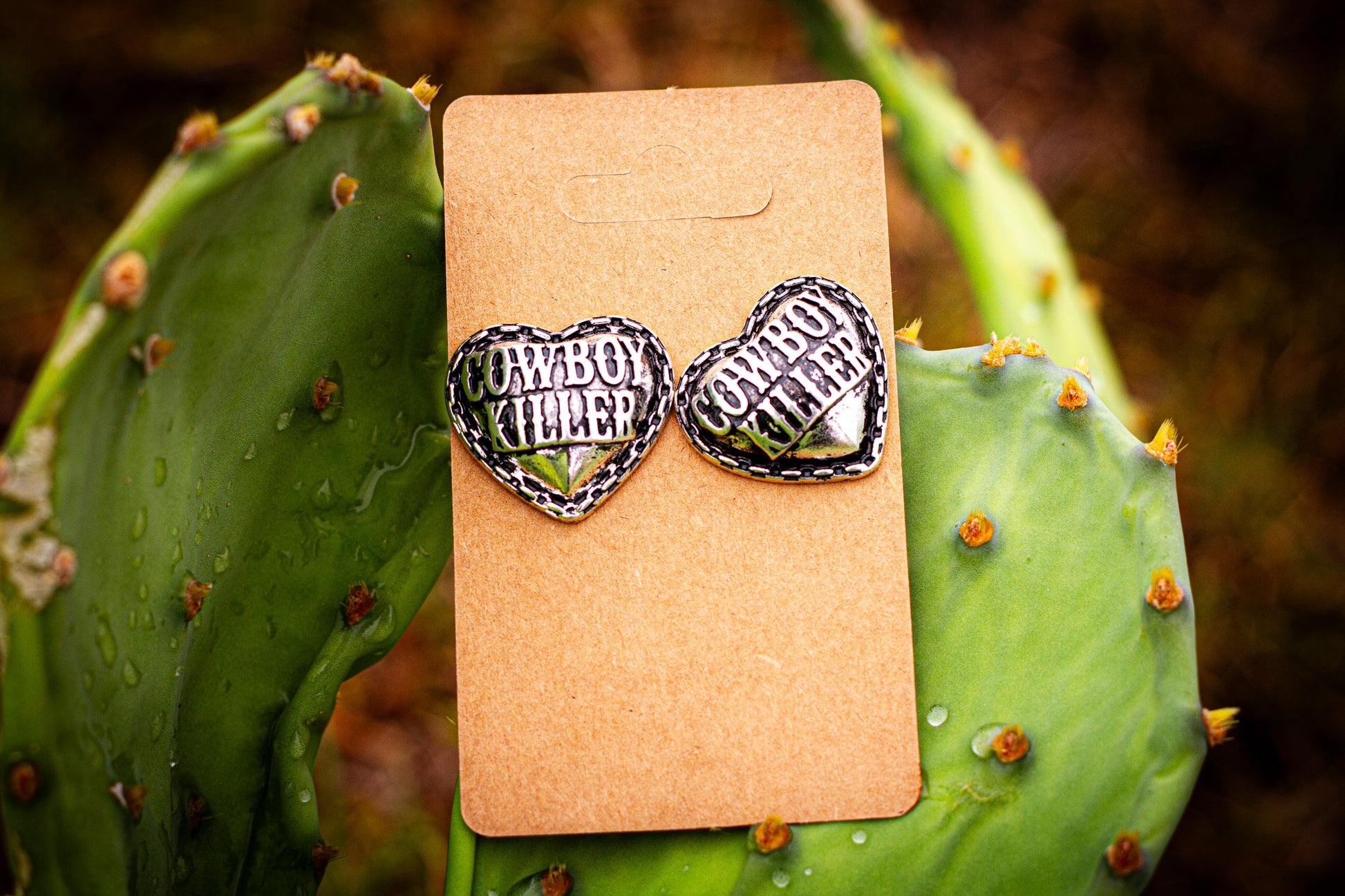 Heart-shaped earrings with 'Cowboy Chiller' text on a cardboard backing, placed on a cactus leaf.