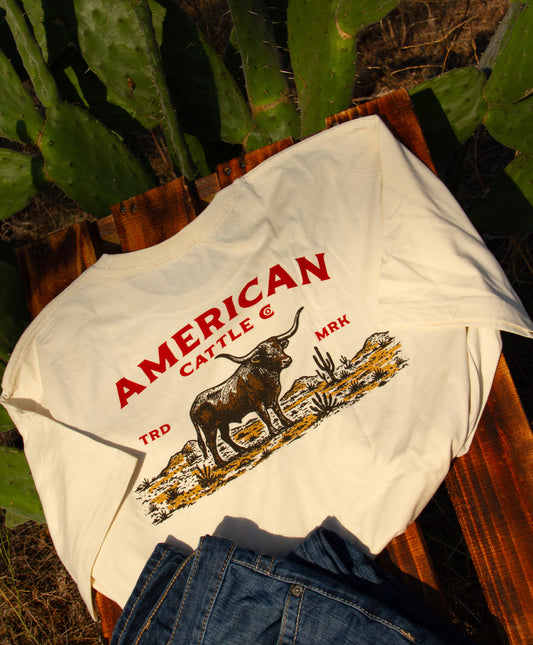 Cream t-shirt with 'American Cattle Co.' logo on a wooden surface with cacti in the background