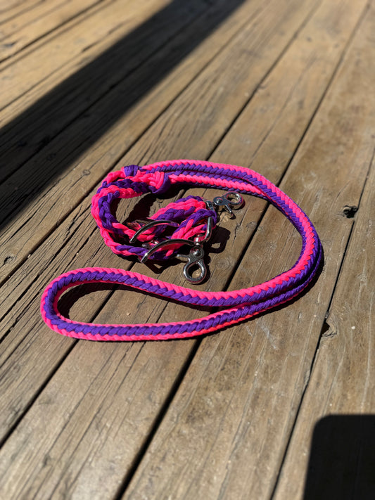 Close-up of hand-braided pink and purple nylon horse reins with trigger snaps, displayed on a wooden surface in natural sunlight.