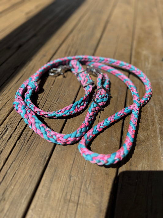 Pink and neon turquoise braided nylon roping reins with stainless trigger snaps and Conway buckles, laid on rustic wooden boards.