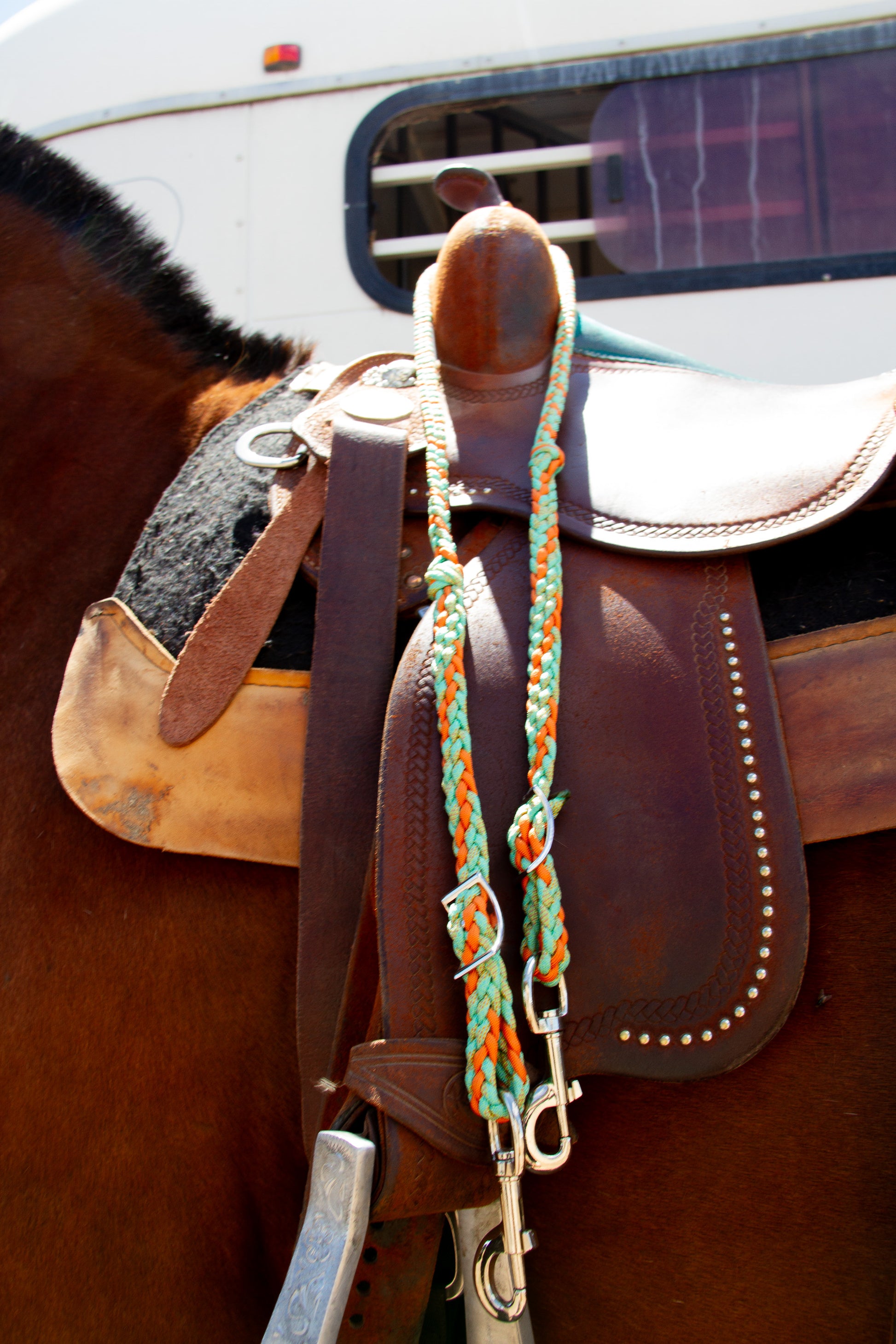 Close-up of a horse saddle with braided reins on a horse, with a vehicle in the background.